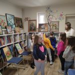 Sala biblioteczna. W białych szafkach stoją poukładane książki. Na nich oraz na krzesłach wystawione są namalowane obrazy z motywami kwiatów, zwierząt oraz martwej natury. Pracownica biblioteki oprowadza grupę dzieci przedszkolnych po wystawie.