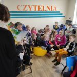 Sala - czytelnia w bibliotece. Dzieci przedszkolne siedzą na krzesłach i żółtej pufie, zwrócone przodem w kierunku pracownicy biblioteki. Kobieta widoczna od tyłu, ubrana w ciemny sweter i biały szal czyta dzieciom książkę. Na białej ścianie widnieje pomarańczowy napis ,, Czytelnia'' oraz namalowane trzy poziome niebieskie pasy.