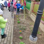 Zdjęcie przedstawia grządkę z posadzonymi kwiatami. Za grządką na chodniku znajdują się dzieci. Część z nich stoi, część siedzi na ławce. W obrębie grządki znajduje się słup latarni i młode drzewko. Po prawej stronie widać dwa przęsła ogrodzenia w kolorach czerwonym i zielonym, po lewej stronie zarysy parkujących aut.