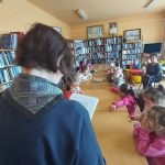 Sześcioro dzieci siedzi w rzędzie przy złączonych stołach i słucha czytania pani bibliotekarki, która trzyma w rękach białe kartki
